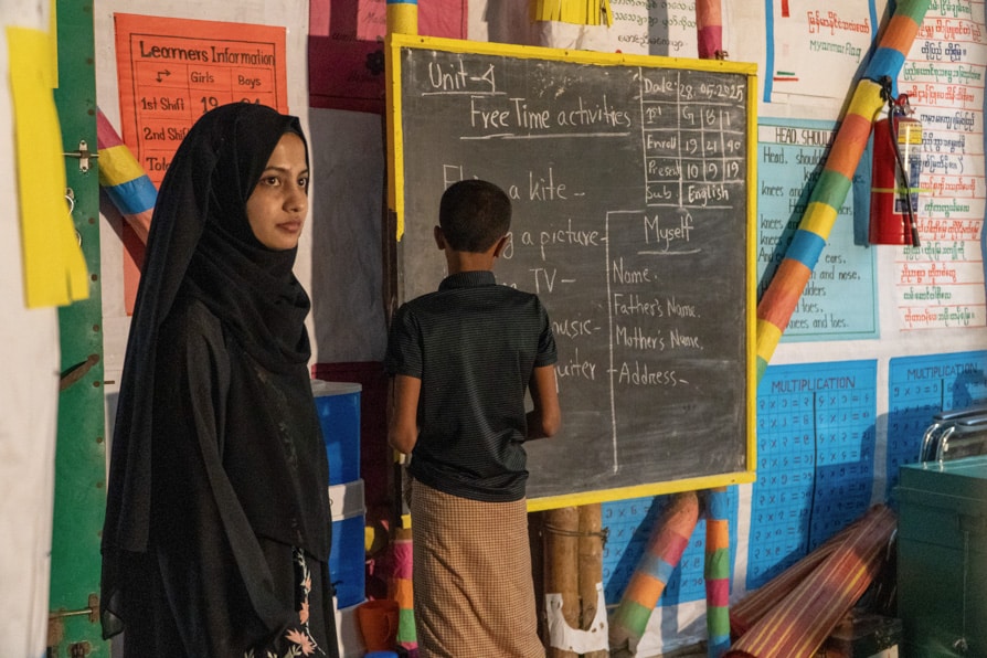 A student writes on a blackboard watched over by a teacher in a learning center in Kutupalong Refugee Camp, Bangladesh.