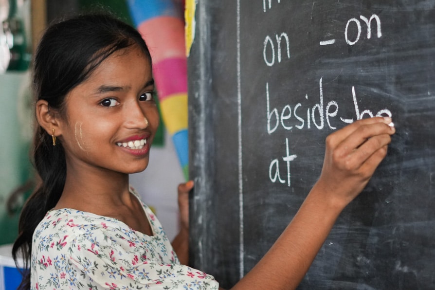 A girl smiles at the camera while writing on the blackboard at a learning center in Kutuplaong Refugee Camp, Bangladesh.