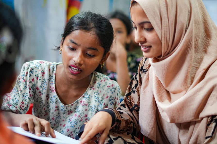 A female teacher wearing a headsccarf helps a female student with her studies during a lesson at a learning center in Kutupalong Refugee Camp, Bangladesh.