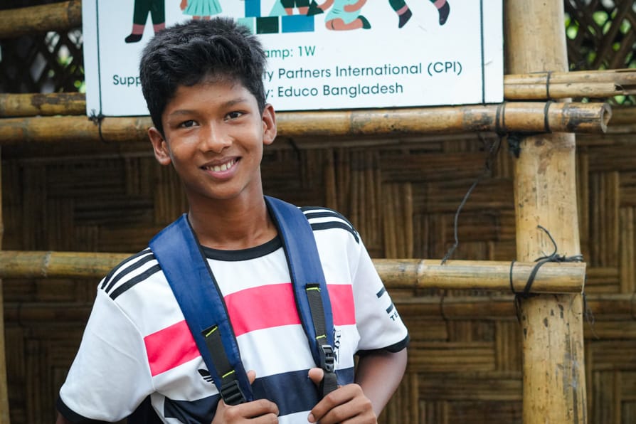 A boy wearing a backpack stands smiling in front of a learnign center in Kutupalong Refugee Camp, Bangaldesh
