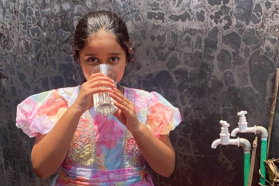 A girl wearing a colorful dress drinks from a glass of water next to a tap that forms part of a safe water network in Kalishakhali village, Bangladesh.