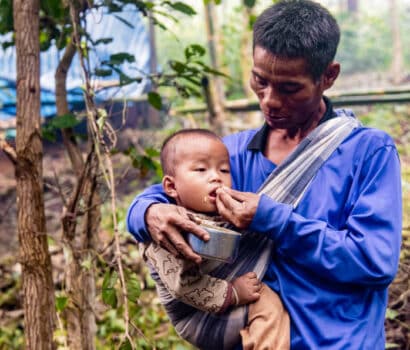 A man holds a child in a sling while feeding him rice at a displacement site in a forest in Myanmar.