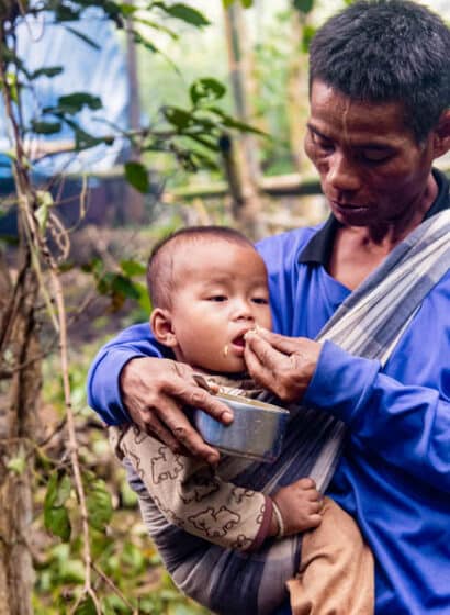 A man holds a child in a sling while feeding him rice at a displacement site in a forest in Myanmar.