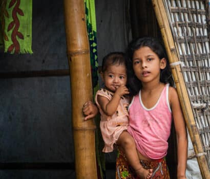 A girl holds her baby brother in the doorway of a bamboo shelter in Kutupalong Refugee Camp, Bangladesh.
