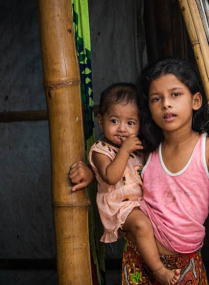 A girl holds her baby brother in the doorway of a bamboo shelter in Kutupalong Refugee Camp, Bangladesh.