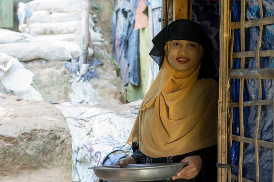 A Rohingya woman stands holding a tray of rice grains at the entrance to her shelter in Kutupalong Refugee Camp, Bangladesh.