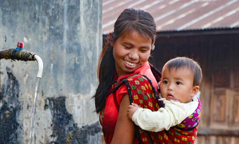 A woman stands smiling with her baby tied to her back in a sling in Myanmar.