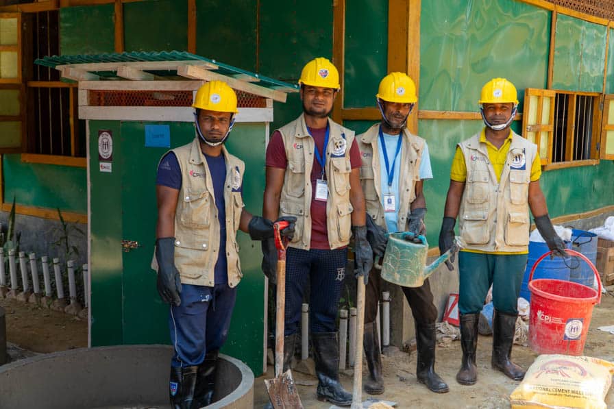 Four Rohingya WASH workers wearing hard hats stand together holding tools in Kutupalong Refugee Camp, Bangladesh.