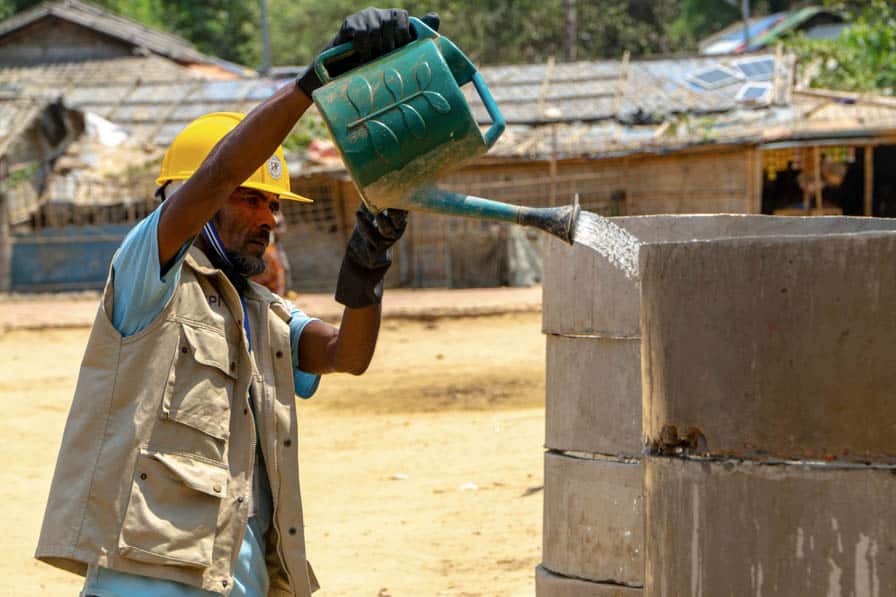 A Rohingya WASH workers pours water from a watering can onto a stack of concrete rings in Kutupalong Refugee Camp, Bangladesh.
